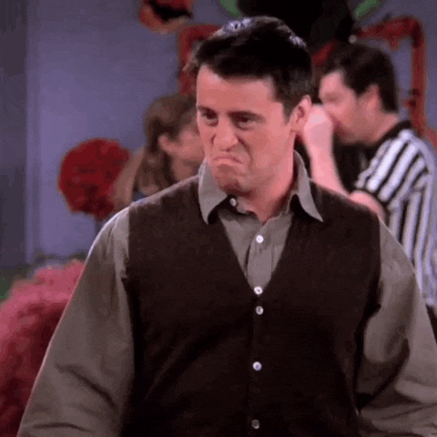 A man in a brown vest and grey shirt looks down and makes an exaggerated frown, showing disappointment or frustration, while people are seen in the background at what appears to be a party.