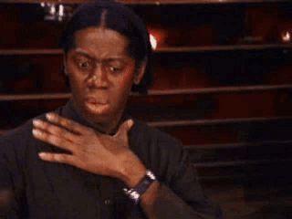 A surprised person wearing a dark shirt holds their chest in shock, with wide eyes and an open mouth, expressing disbelief or astonishment.