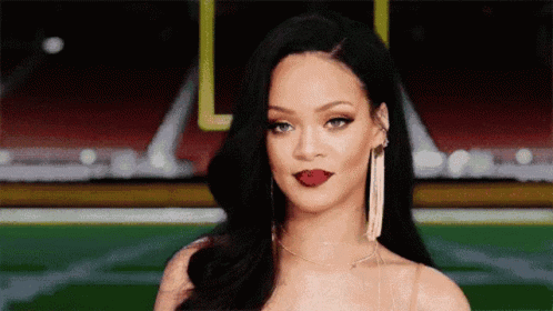 A woman with long dark hair and bold red lipstick gives a subtle smirk while standing in a stadium. She is wearing long statement earrings, and her expression exudes confidence.