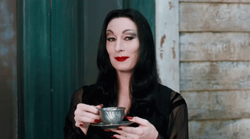 A woman with long dark hair, wearing a black outfit and red lipstick, elegantly sips tea from a cup while raising her eyebrows in a sly, knowing expression.